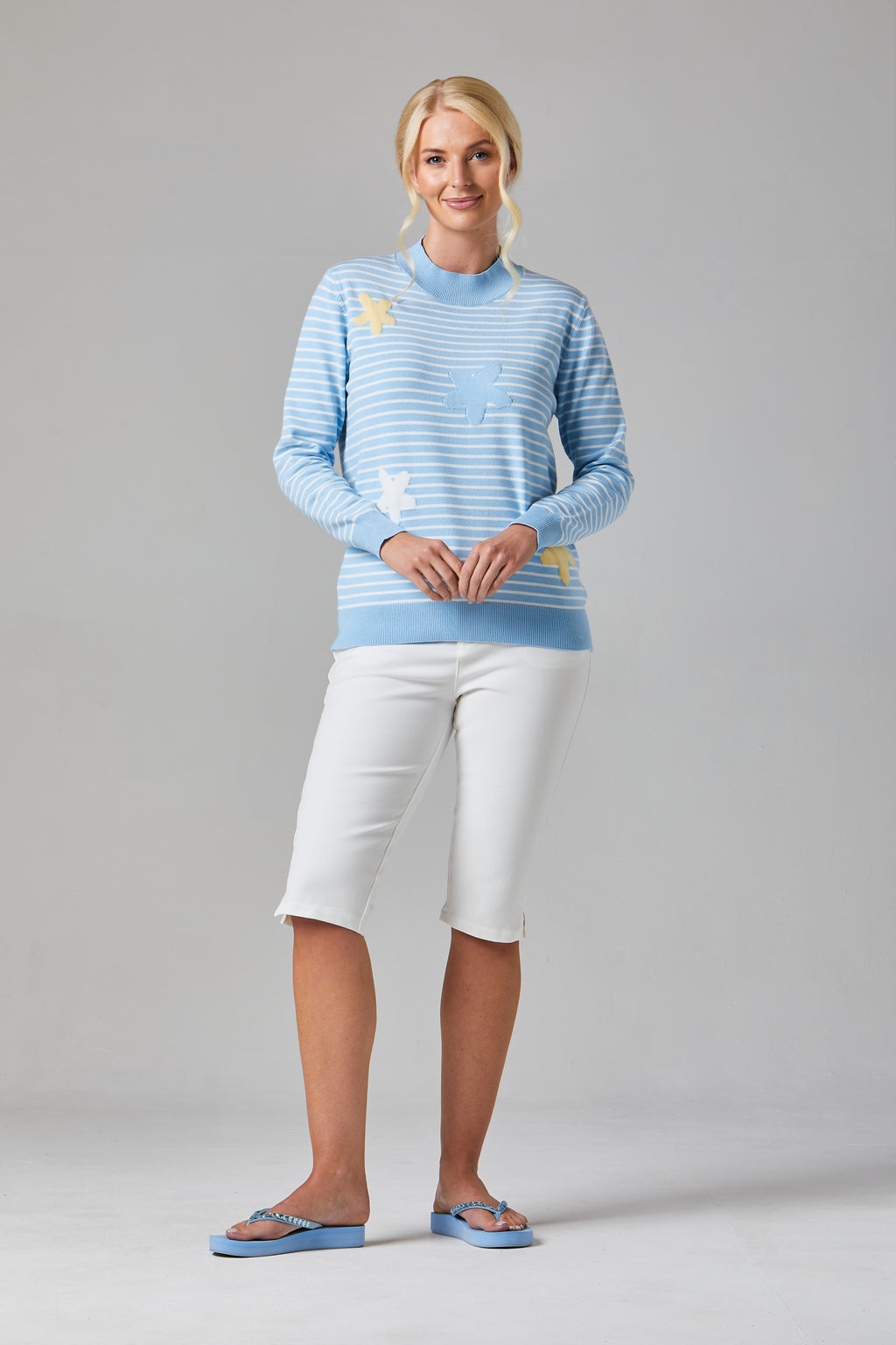 Buy Online Petula Pullover Alice Collins -  Woman wearing a light blue striped sweater and white pants on a gray background
