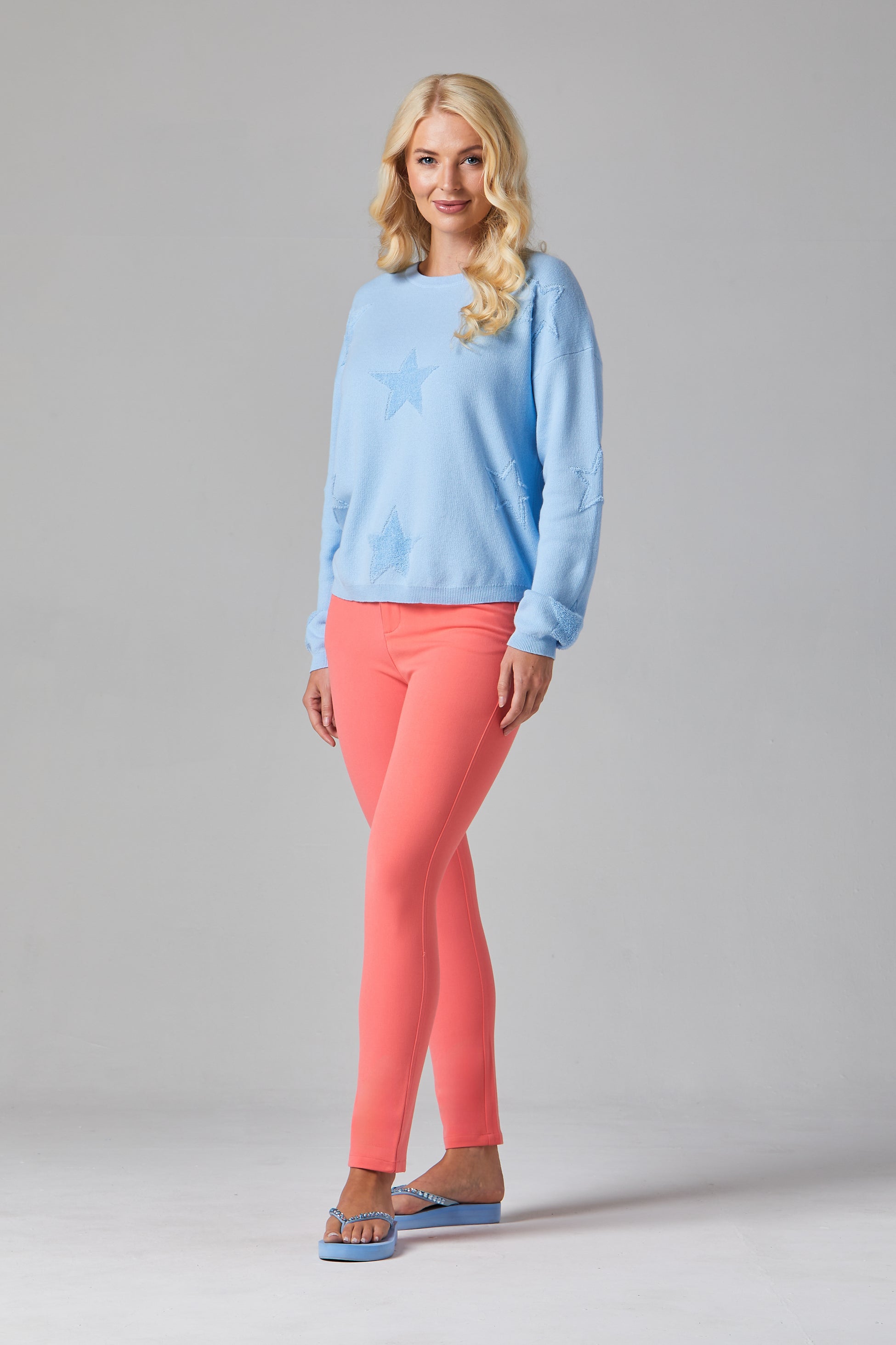 Buy Online Mimi Pullover Alice Collins Woman wearing a light blue sweater and pink pants on a gray background