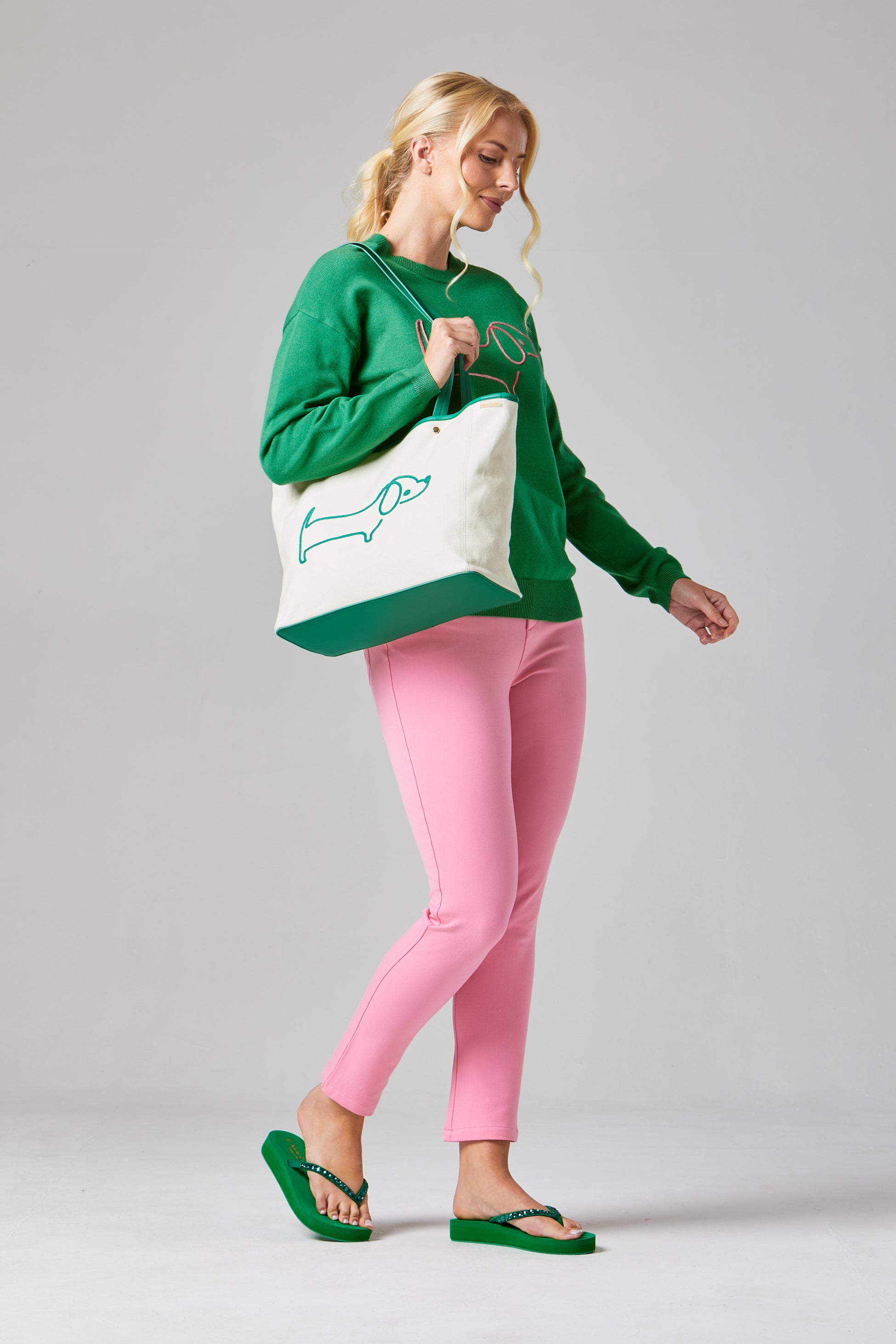 Buy Online Sausage Dog Pullover Alice Collins -Woman holding a tote bag with a green design, wearing a green sweatshirt and pink pants on a gray background