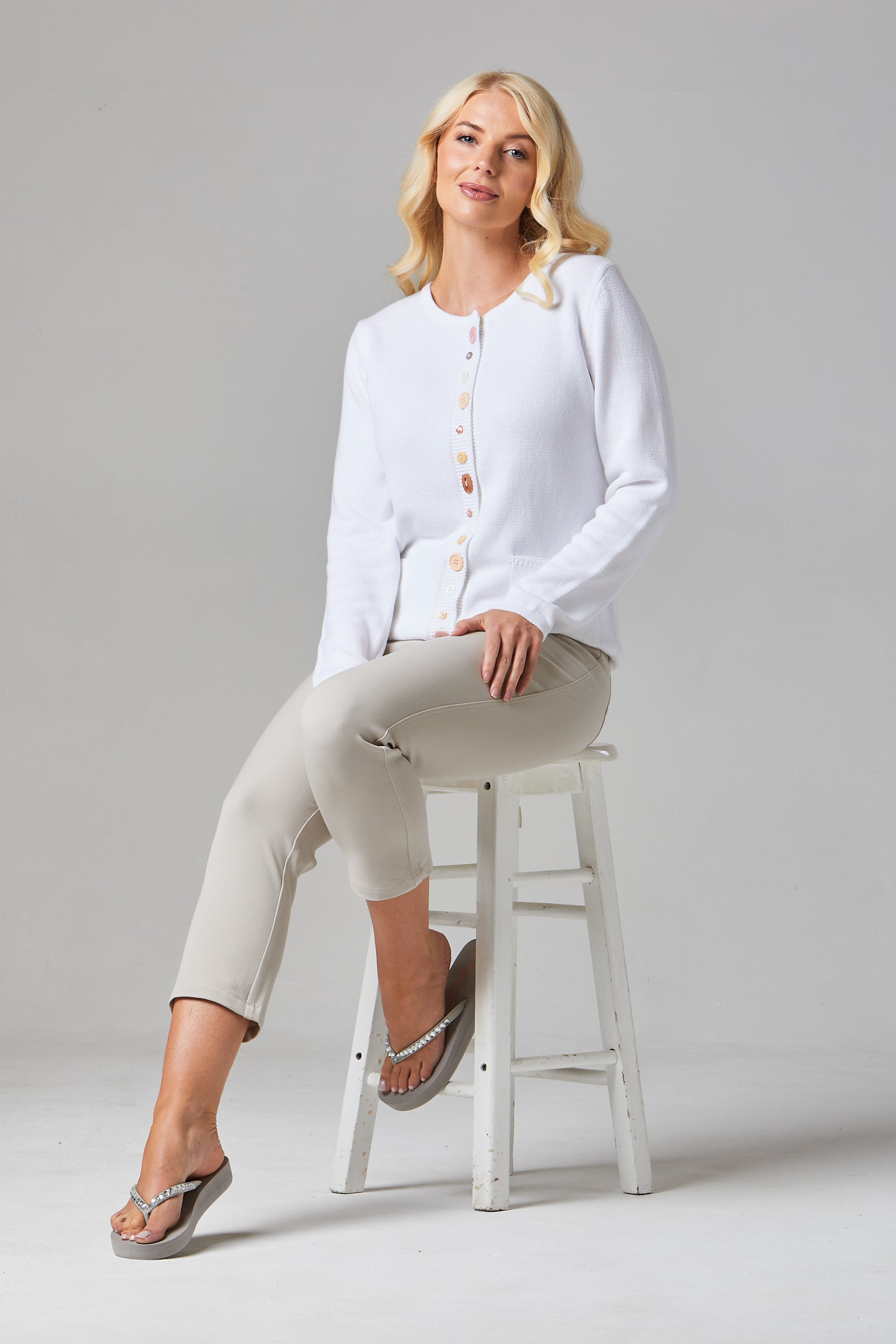 Buy Multi Button Cardigan Online - Alice Collins - Woman sitting on a stool wearing a white cardigan and beige pants against a gray background